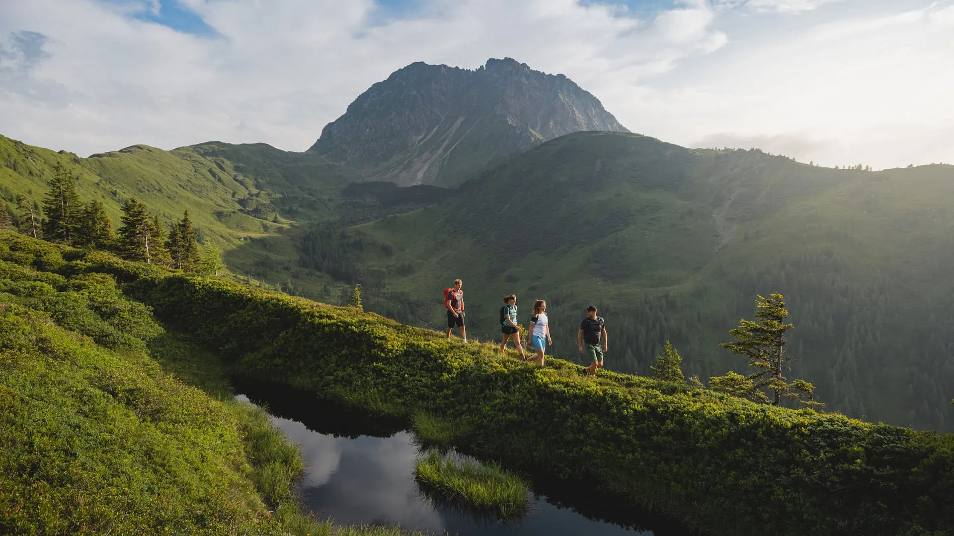 Gruppe von Menschen wandert auf einem grünen Hügel mit Bergen im Hintergrund.