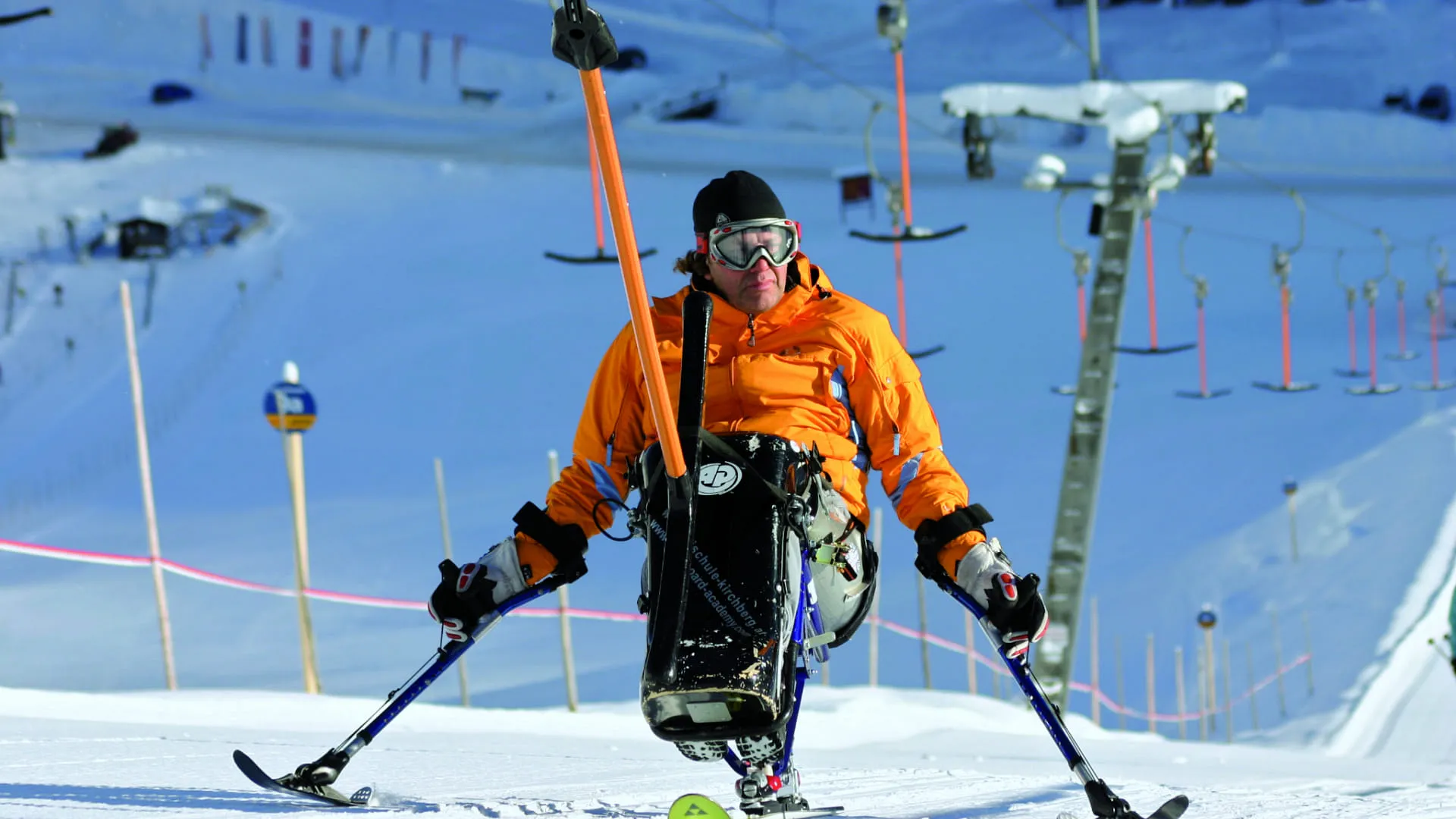 Skifahrer in orangeem Anzug auf schneebedeckter Piste mit Skistöcken